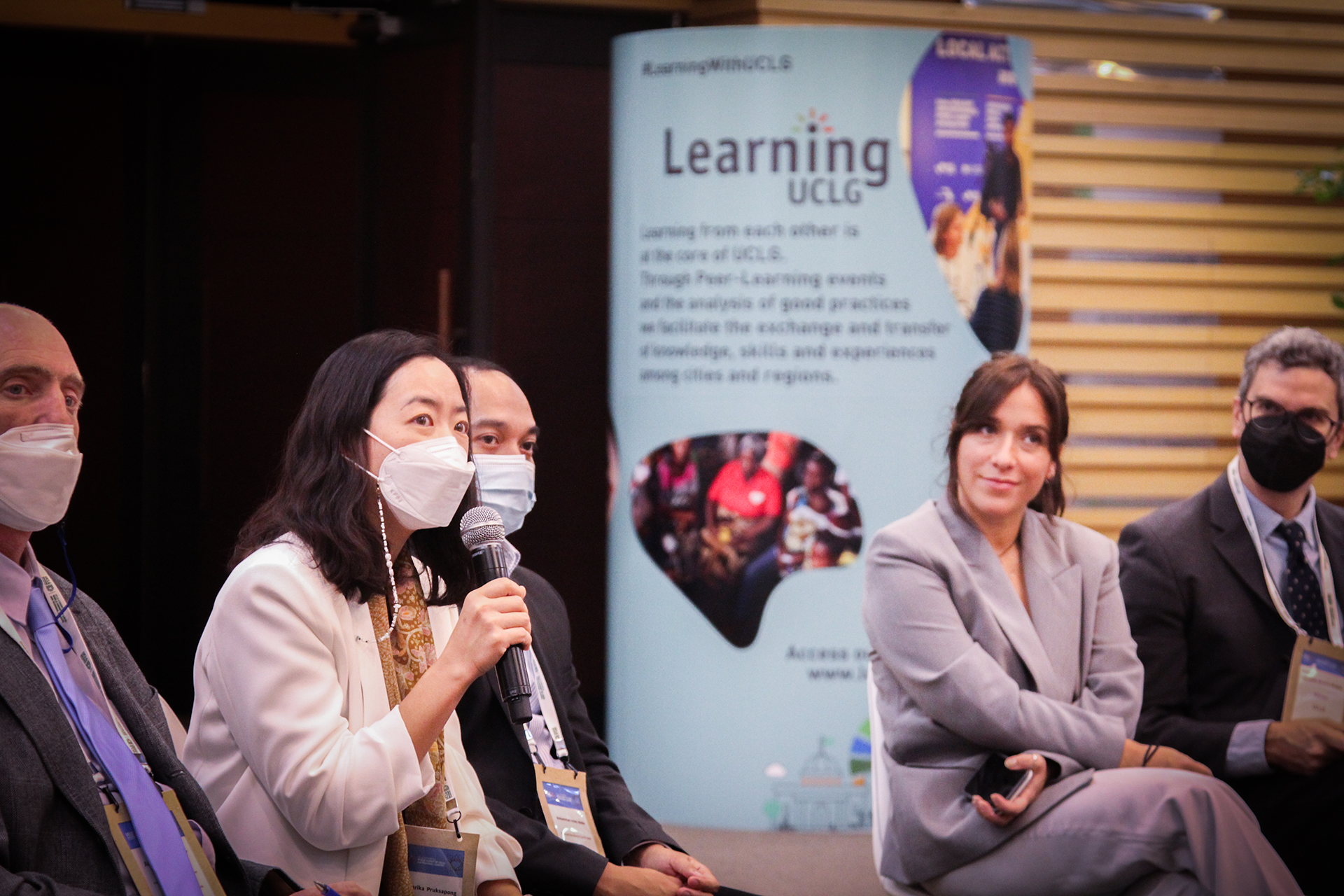 1.	Una mujer con mascarilla sostiene un micrófono mientras está sentada entre otras personas. Una columna con la identidad de CGLU Learning aparece en el fondo.