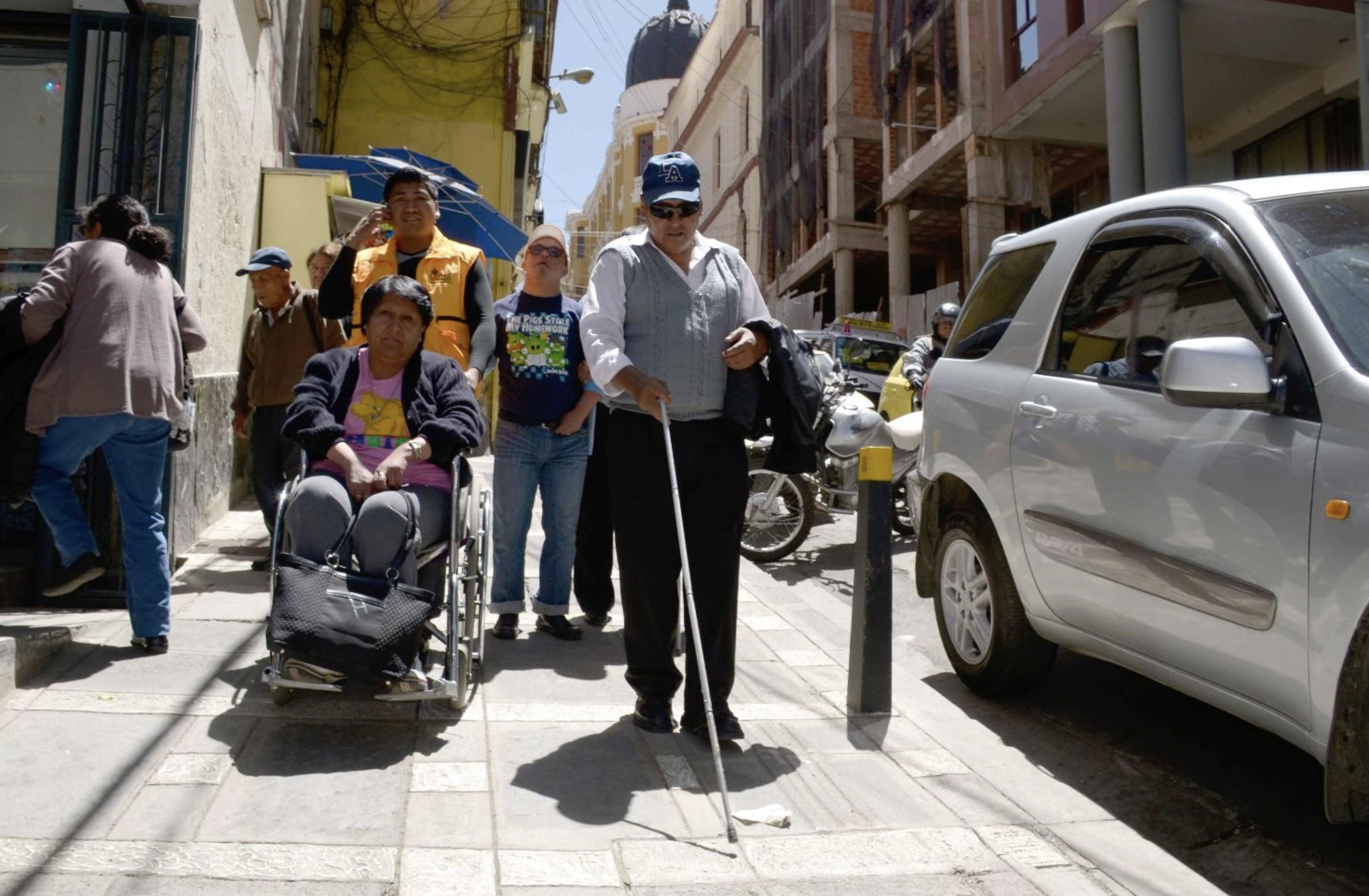 Diverses personnes se déplacent dans la ville avec différentes aides à la mobilité, notamment des cannes blanches et des fauteuils roulants.